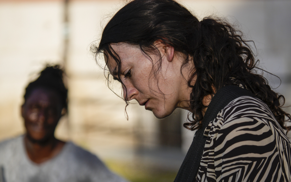 Teresa is a white woman with long brown hair tied back in a ponytail. She wears a black and white striped long-sleeved blouse. She plays a red guitar with a black neck strap. A dark-skinned second musician sits in the background and looks over at her.