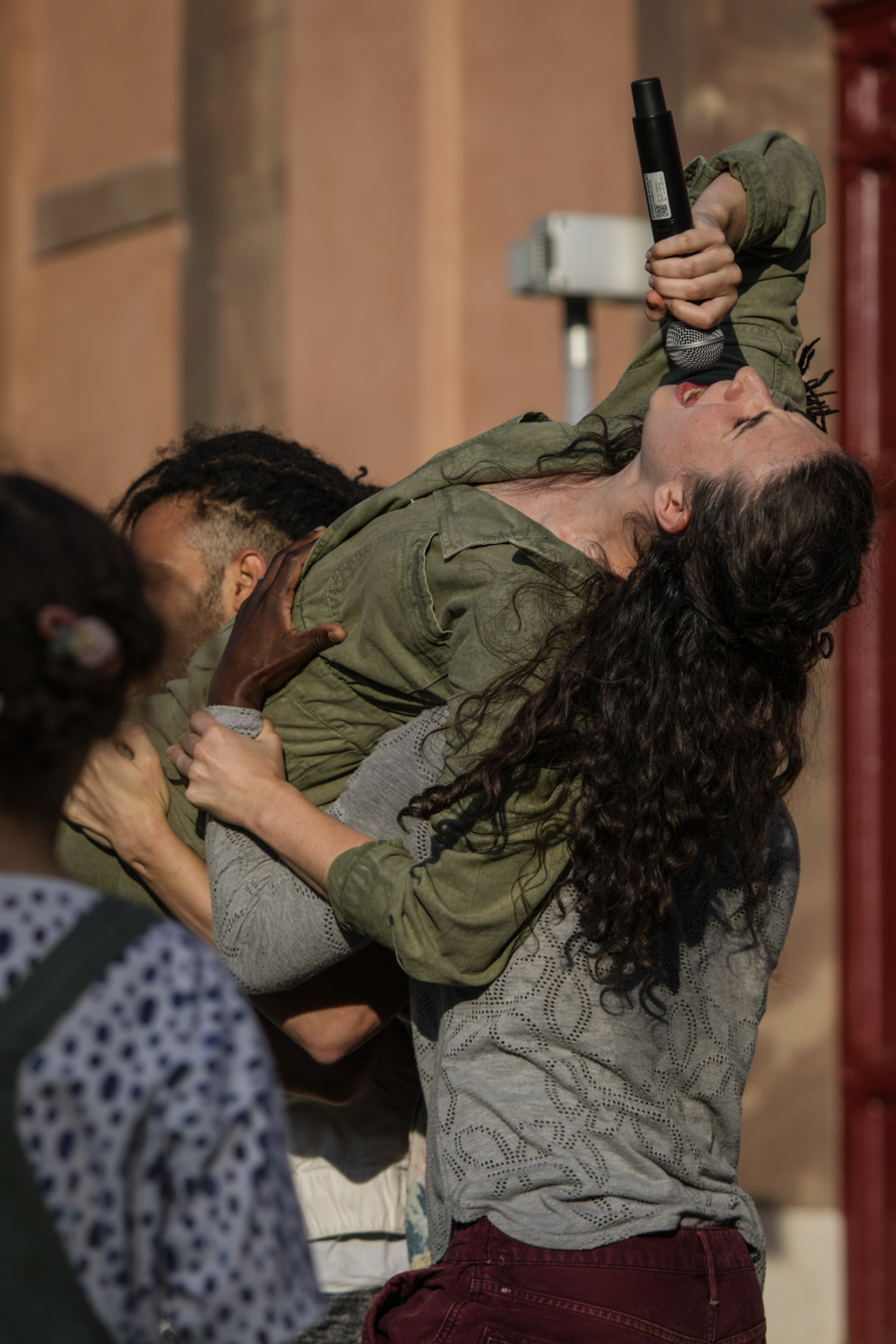 Katy is a white woman with long brown hair that falls down her back. She is wearing an olive green long-sleeved jumpsuit. Her head is tilted back and she sings energetically into a microphone she holds in her right hand while being carried on her shoulders by three other dancers. The wall of a sandstone-coloured house can be seen in the background.