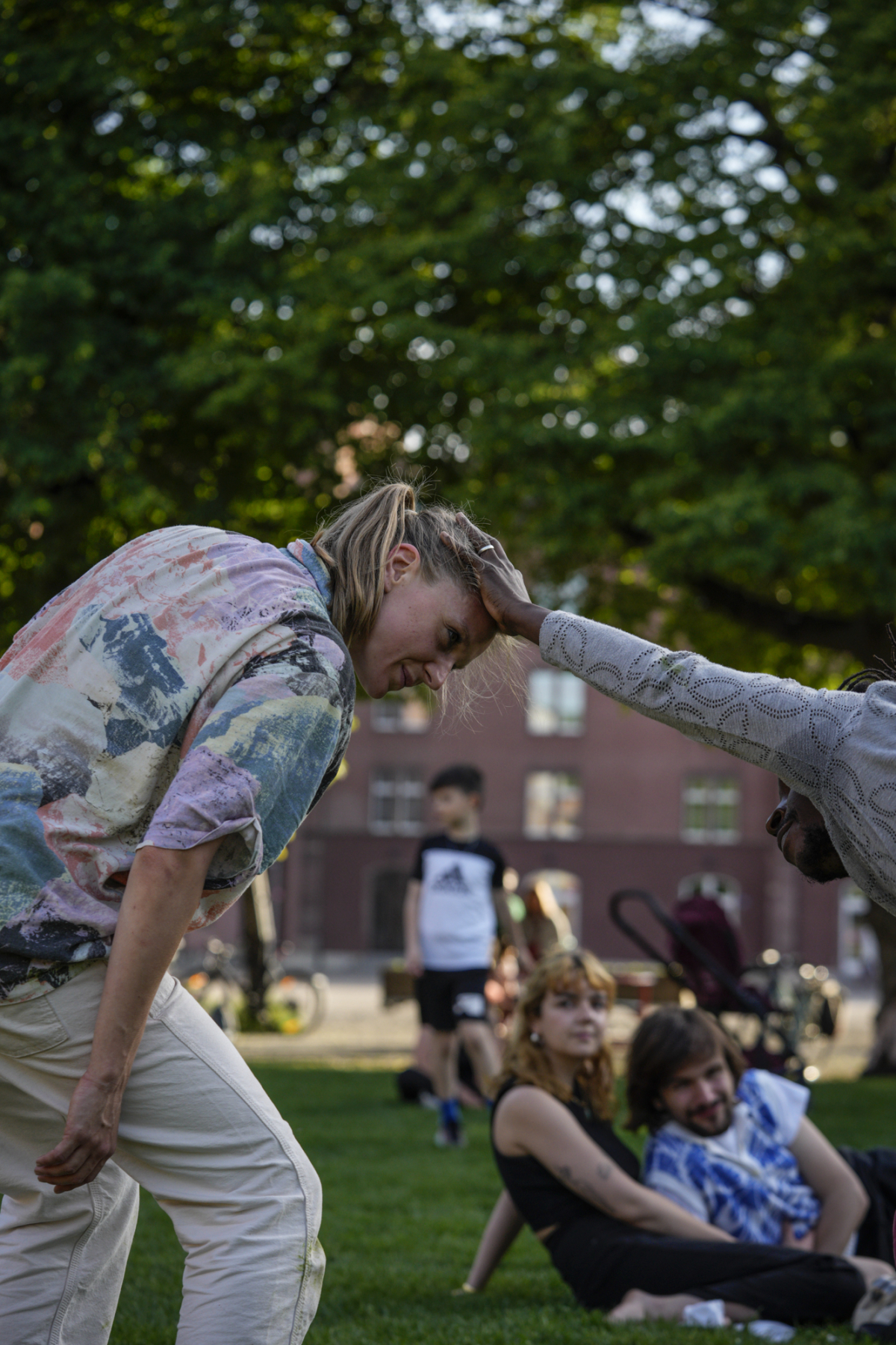 Melissa is a white woman with long blonde hair tied back in a ponytail. She is wearing a colourful shirt and white trousers. She smiles as she bends her knees and tilts her head and face forward towards another dancer who smiles at her and holds her head with his outstretched right hand. They are in a park with green grass and a green lush tree in the background and an audience watching from close by.