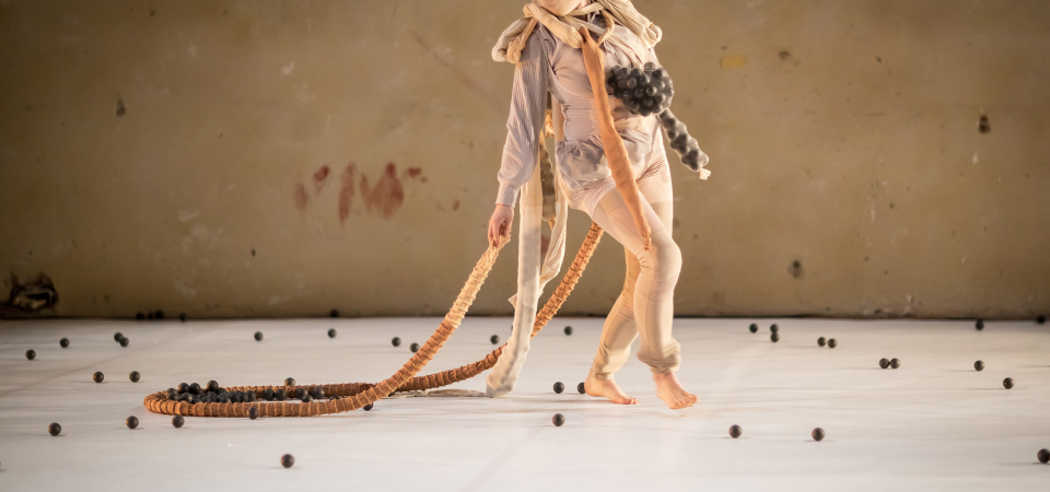 Eine Figur mit schwarzer Perücke, gekleidet in ein helles, dehnbares Tuch, läuft über eine Bühne, die mit kleinen schwarzen Bällen übersät ist. Launig zieht sie einen kleinen Haufen der schwarzen Bälle hinter sich her.